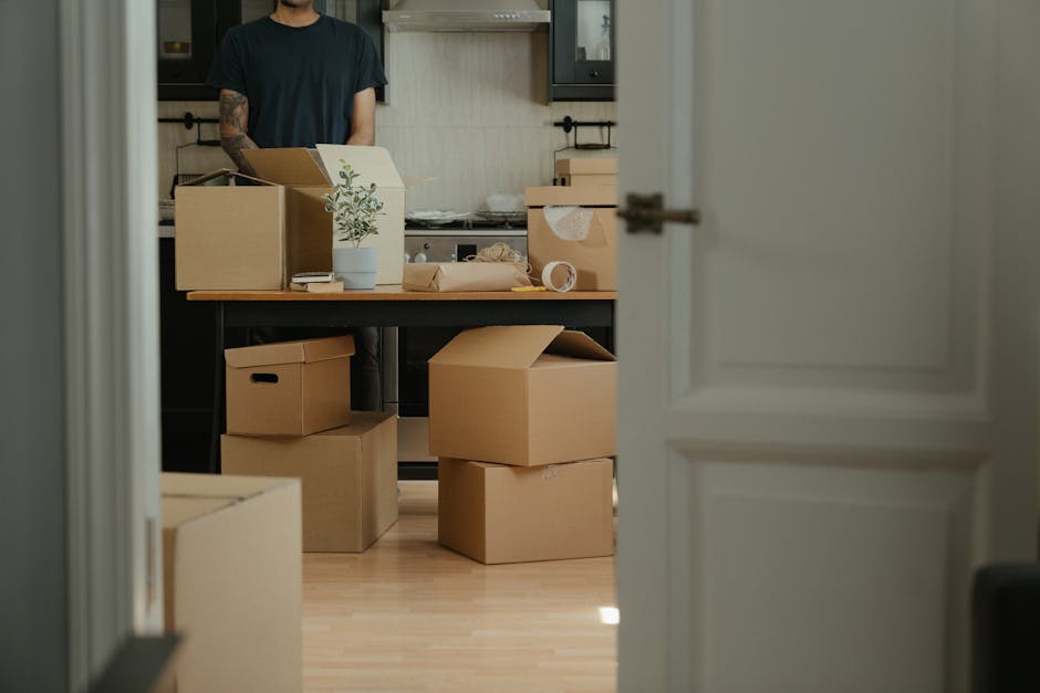 A view through a partially open door into a room being prepared for home relocation, showing a black dining table covered with packing materials and cardboard boxes. Several cardboard boxes of various sizes, some sealed and others open, are stacked on the floor and on the table, with brown packing paper, plastic wrap, and tape visible around them. A small potted plant is also placed on the table among the packing supplies. In the background, a person dressed in a black T-shirt is seen standing behind the table, possibly organising or packing items, with a modern kitchen visible on the back wall featuring dark cabinets and a light countertop. The scene indicates an active packing and furniture transport process typical of house removals, with the environment well-lit by natural light, and the use of packing boxes, wrapping materials, and furniture moving equipment reflects professional moving services provided by Man and Van Aldersbrook, aligning with the services highlighted in the Aldersbrook removals guide for Wanstead Flats moves.