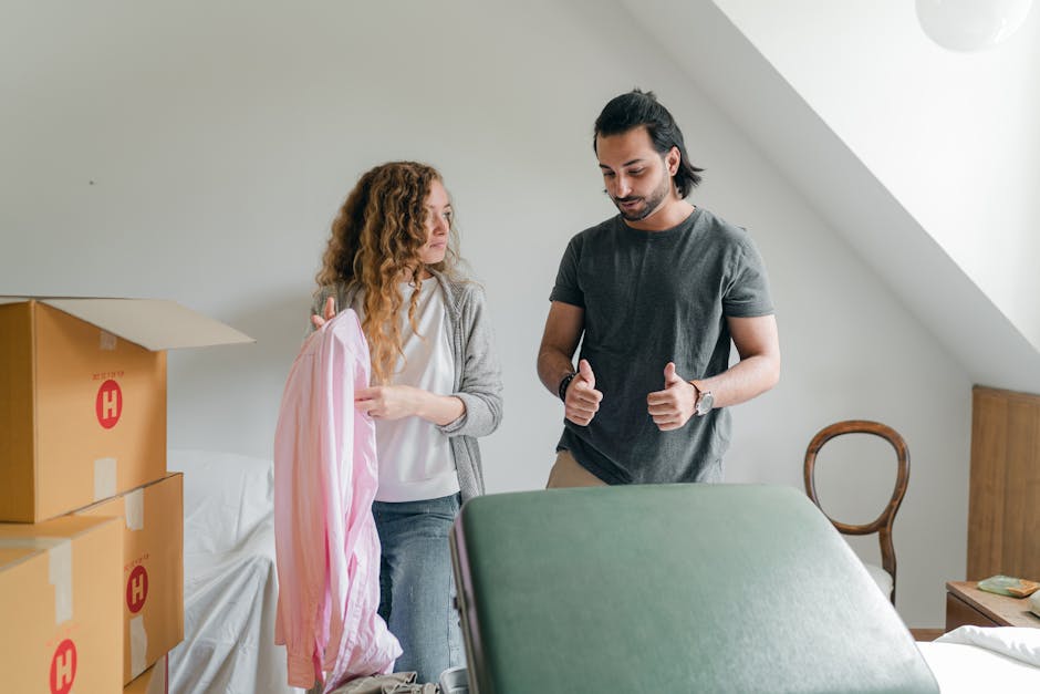 A man and a woman are indoors, preparing for a house removal. The woman, with curly red hair, is holding a pink garment bag with clothing inside and appears to be discussing the moving process. The man, with dark hair and a beard, is giving a thumbs-up gesture and looks focused. The room has a plain white wall and natural lighting coming from an unseen window. In the background, there are cardboard boxes with red markings stacked on a white sofa, which are likely packed for moving. The boxes and garment bag suggest packing and relocation preparations are underway. A green moving blanket is draped over a piece of furniture, possibly a table or dresser, in the foreground, indicating furniture protection during transport. A wooden chair is positioned against the wall, and a small side table with a decorative item is visible. The scene depicts a typical preparation phase for home relocation, involving packing clothes, securing furniture, and organizing boxes for transport, consistent with services offered by Man and Van Aldersbrook during furniture transport and removal processes.