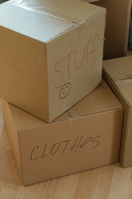 A view through a partially open door into a room being prepared for home relocation, showing a black dining table covered with packing materials and cardboard boxes. Several cardboard boxes of various sizes, some sealed and others open, are stacked on the floor and on the table, with brown packing paper, plastic wrap, and tape visible around them. A small potted plant is also placed on the table among the packing supplies. In the background, a person dressed in a black T-shirt is seen standing behind the table, possibly organising or packing items, with a modern kitchen visible on the back wall featuring dark cabinets and a light countertop. The scene indicates an active packing and furniture transport process typical of house removals, with the environment well-lit by natural light, and the use of packing boxes, wrapping materials, and furniture moving equipment reflects professional moving services provided by Man and Van Aldersbrook, aligning with the services highlighted in the Aldersbrook removals guide for Wanstead Flats moves.