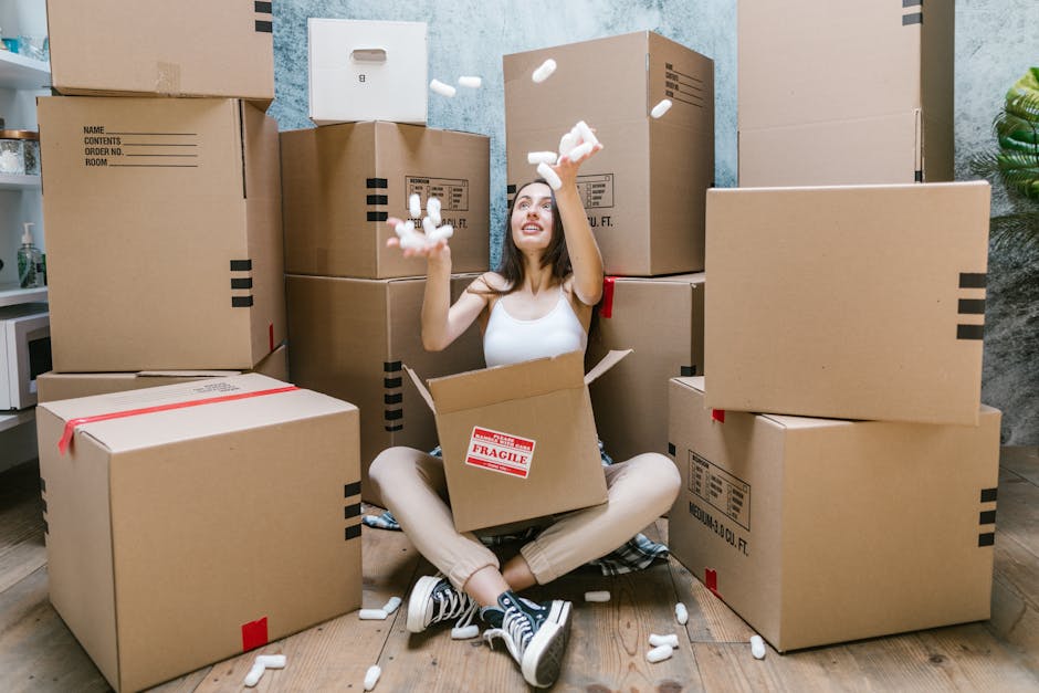 A young woman sitting cross-legged on a wooden floor surrounded by packed cardboard boxes with black and red labels, some sealed with red tape, during a home relocation process. She is smiling and tossing foam packing peanuts into the air, with several peanuts also falling around her. The boxes vary in size and are stacked against a blue and grey textured wall, indicating an interior setting prepared for a move. She is wearing casual clothing, including a white top and black sneakers, and appears to be in a preparation stage for a moving service offered by Man and Van Aldersbrook. A cardboard box sitting on her lap has a 'Fragile' sticker. A glimpse of a white appliance or furniture piece is visible on the left, and part of a potted plant can be seen on the right, suggesting an organized packing environment in readiness for furniture transport or house removals as part of the moving process.