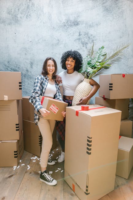 Two women are standing inside a room filled with several cardboard moving boxes, some sealed with red tape and others partially open, containing packing materials such as white paper or foam packing peanuts. The woman on the left is holding a medium-sized cardboard box, dressed in beige pants, black and white sneakers, a plaid shirt over a white top, and has shoulder-length brown hair. The woman on the right is holding a large, decorative woven vase with green palm leaves poking out, and is wearing patterned pants, a white top, with curly black hair. They are smiling, standing close together, indicating a positive moment during a home relocation process. The environment appears to be a transitional space, possibly a living room or storage area, with a plain light blue or grey textured wall behind them and a wooden floor underneath. The scene captures part of a furniture move or packing activity, with visible movement and items awaiting transportation, consistent with the services provided by Man and Van Aldersbrook for same-day local moves involved in furniture transport and loading processes.