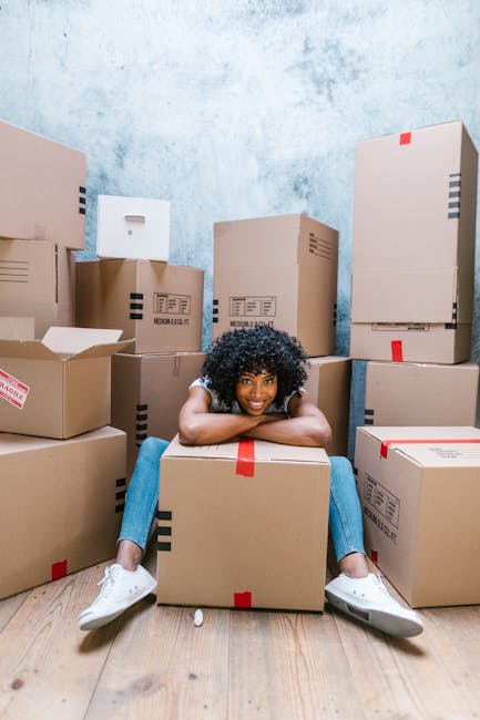 A young woman with curly black hair, wearing a white t-shirt, blue jeans, and white sneakers, is sitting on a wooden floor inside a room filled with stacked cardboard boxes of various sizes. Some boxes are sealed with red packing tape, while others are partially open revealing packing paper inside. The woman is smiling and resting her chin on her folded arms atop a medium-sized box. The background features a plain, lightly textured light blue wall, and the scene appears to depict a home relocation or packing process underway, with boxes arranged around her as part of a furniture transport or moving preparation, associated with services offered by Man and Van Aldersbrook.