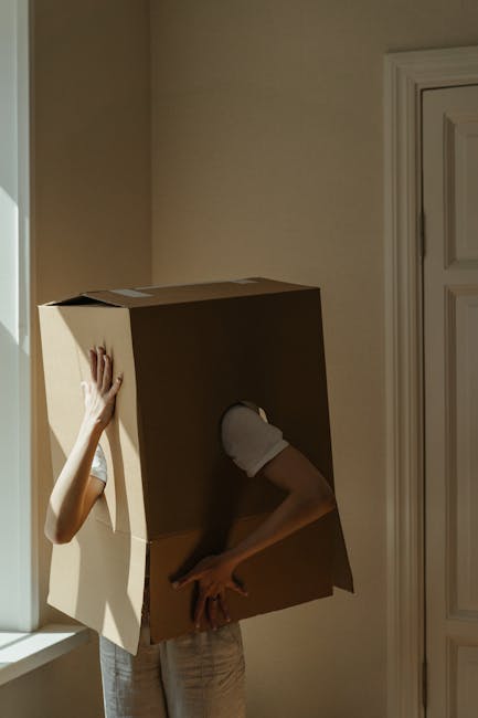 A person wearing white gloves is lifting a large cardboard moving box, which is placed over their head and shoulders, inside a room with beige walls and a white window frame. The individual is preparing to load the box onto a transport vehicle as part of a home relocation process. Visible in the background are other cardboard boxes and packing materials, indicating an active packing and moving scene. The lighting is natural, coming from the window nearby, and the room appears tidy, supporting a professional furniture transport and packing operation conducted by Man and Van Aldersbrook.
