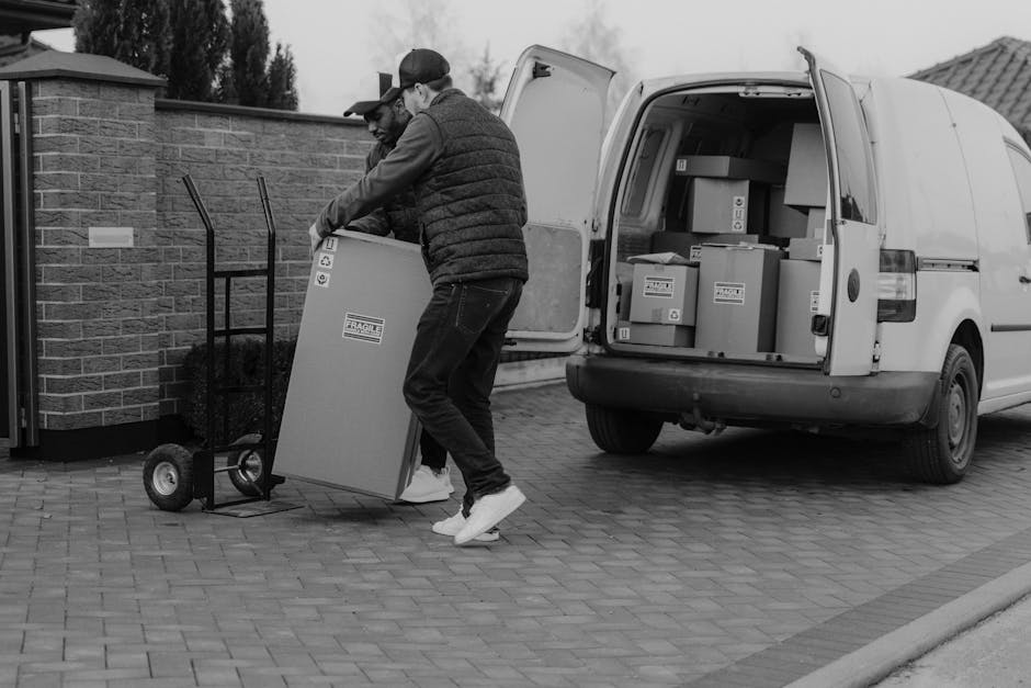 A man wearing a yellow padded vest, grey jeans, and a black cap, is opening the sliding side door of a white commercial van parked on a driveway made of brick pavers. Inside the van, several cardboard
