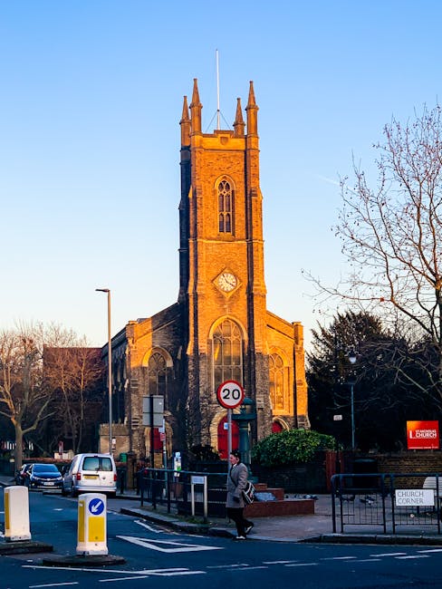 A tall, historic stone church with a square tower featuring pointed pinnacles and narrow arched windows, situated on a street corner during daylight with a clear blue sky. In the foreground, a person