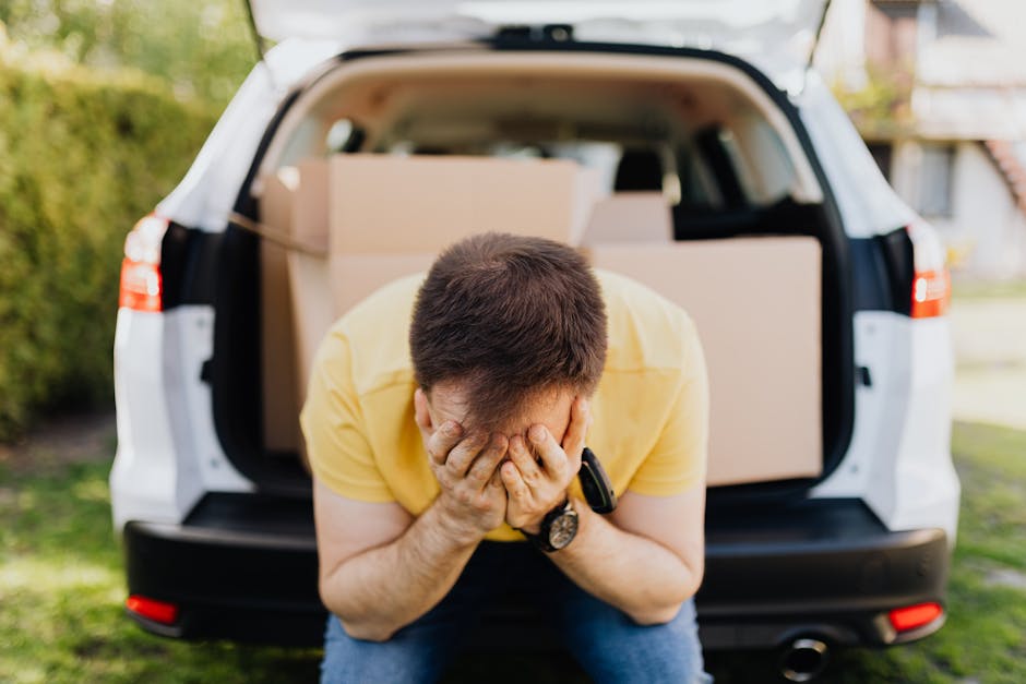 A man wearing a yellow shirt sits on a transport vehicle's open tailgate with his head in his hands, displaying frustration or stress. Behind him, inside the vehicle's cargo area, several cardboard boxes are visible, some wrapped in protective plastic film. The boxes are positioned on the vehicle's interior floor, with larger boxes towards the back and smaller ones in front. The vehicle is parked outdoors on a grassy area, with trees and residential houses in the background, suggesting a home relocation setting. The scene captures a moment of handling packing or loading challenges during furniture transport, aligning with the context of home removals and last-minute bookings in response to delays, as addressed by Man and Van Aldersbrook.