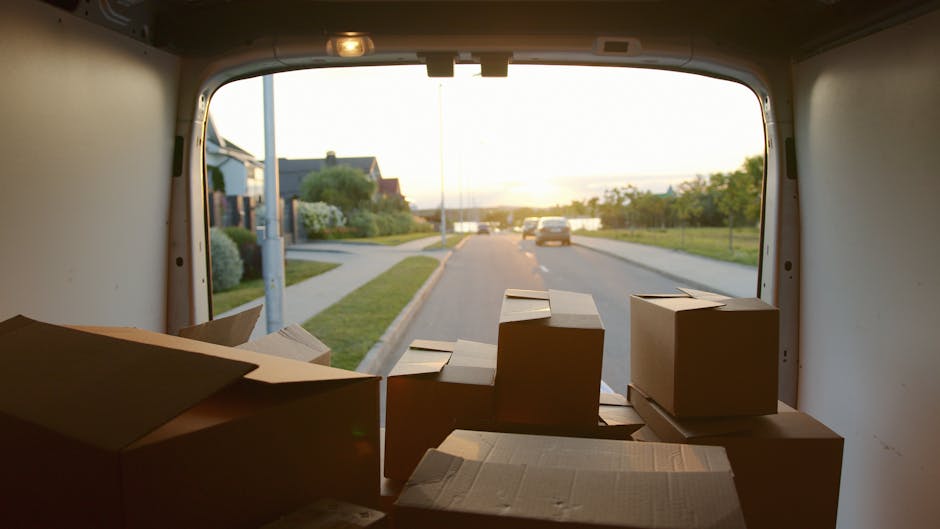View from inside a moving van showing several cardboard boxes placed on the floor near the rear opening, ready for loading or unloading during a home relocation. The boxes vary in size and are sealed with packing tape, with some partially open revealing paper or packaging material inside. The van's spacious interior is illuminated by natural light, and the scene captures a suburban street outside with parked cars, houses, greenery, and a sunset sky, indicating late afternoon or early evening. The environment suggests a professional furniture transport process as part of last-minute house removal arrangements. Man and Van Aldersbrook's removal service is depicted through the organized loading setup, supporting efficient packing and moving operations.