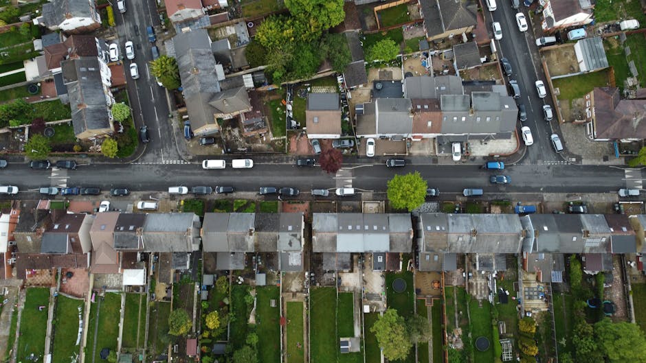 Aerial view of a residential street with closely packed terraced houses and individual backyards, showing parked cars along the curb and behind the houses. The street is lined with driveways, some with cars parked on them, while others have small gardens or paved areas. Trees and greenery are visible in the front and backyards, providing a suburban setting. The image captures a typical urban neighbourhood environment, with a variety of house styles, roofing materials, and landscaping. It illustrates the type of area where house removals and furniture transport services by Man and Van Aldersbrook may operate during home relocation projects, particularly along busy streets with constrained parking and access points.