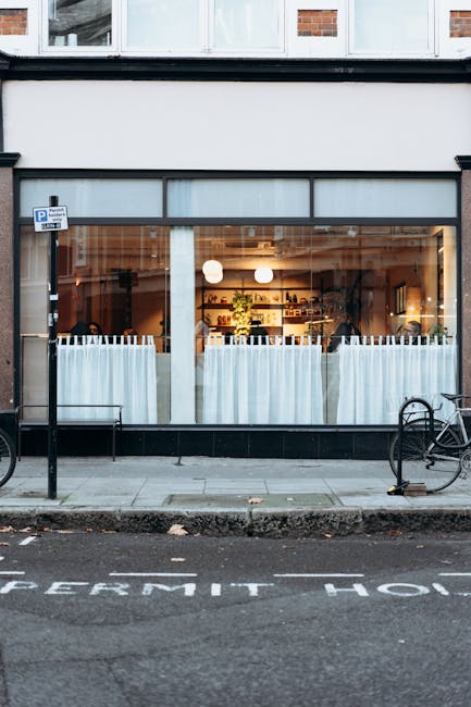 The image shows the front exterior of a commercial premises with large glass windows and a door, revealing the interior illuminated by warm lighting and furnished with shelves and decorative plants. Outside, the pavement includes a metal railing and a bicycle locked to a bike rack on the right. The pavement features a painted sign indicating parking restrictions, including the words 'PERMIT H’ visible across the street. In the foreground, a moving process is taking place; several cardboard boxes and wrapped furniture items are being loaded into a black cargo van parked directly in front of the building. The van’s rear doors are open, and a person (not fully visible) is engaged in lifting or placing items inside. The exterior environment suggests a busy street scene, suitable for home or office relocation activities, with Man and Van Aldersbrook providing professional removals and furniture transport services according to the context of permits for Aldersbrook house moves on busy streets.