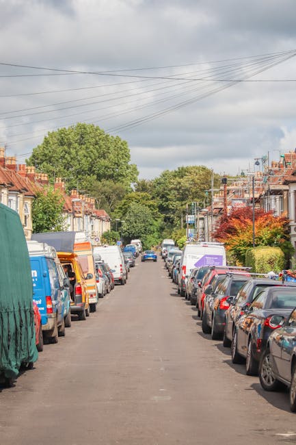 A residential street scene showing a narrow paved road lined with parked cars on both sides, including compact and larger vehicles in various colours. Some vehicles are covered with protective tarpaulins or plastic wraps. The street is flanked by terraced houses with brick facades, small front gardens, and hedges or trees. In the foreground, a moving van from Man and Van Aldersbrook is positioned on the pavement near the curb, with its rear doors open and a hand truck nearby, indicating active loading or unloading. Nearby, several large cardboard boxes, wrapped furniture, and packing materials are visible, suggesting a home relocation process. The surrounding environment is shaded under an overcast sky, with overhead power lines and street lighting fixtures visible. The scene captures the typical logistics involved in house removals, with a focus on the careful transportation of furniture and belongings during a move, as facilitated by professional movers specializing in local removals in Aldersbrook.