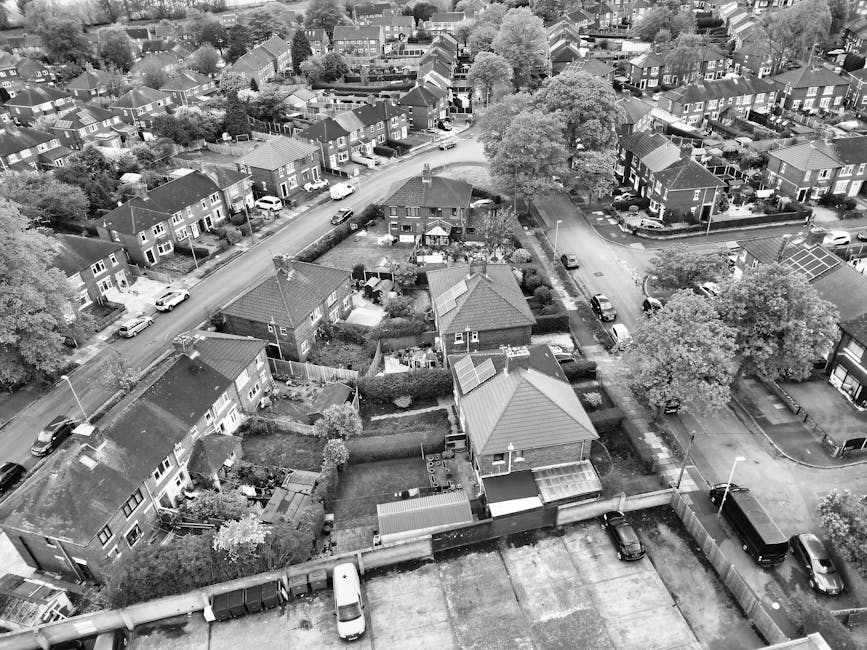 A residential street scene showing a narrow paved road lined with parked cars on both sides, including compact and larger vehicles in various colours. Some vehicles are covered with protective tarpaulins or plastic wraps. The street is flanked by terraced houses with brick facades, small front gardens, and hedges or trees. In the foreground, a moving van from Man and Van Aldersbrook is positioned on the pavement near the curb, with its rear doors open and a hand truck nearby, indicating active loading or unloading. Nearby, several large cardboard boxes, wrapped furniture, and packing materials are visible, suggesting a home relocation process. The surrounding environment is shaded under an overcast sky, with overhead power lines and street lighting fixtures visible. The scene captures the typical logistics involved in house removals, with a focus on the careful transportation of furniture and belongings during a move, as facilitated by professional movers specializing in local removals in Aldersbrook.
