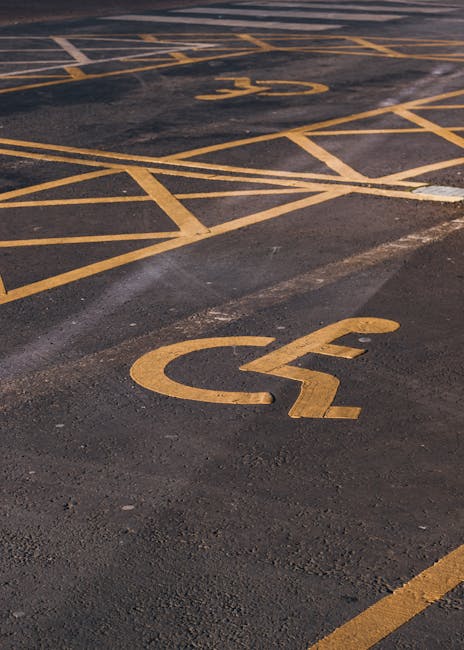 Close-up view of a parking area pavement with yellow lines and markings designated for disabled parking spaces, including a large wheelchair symbol painted on the asphalt. The surface appears clean wi