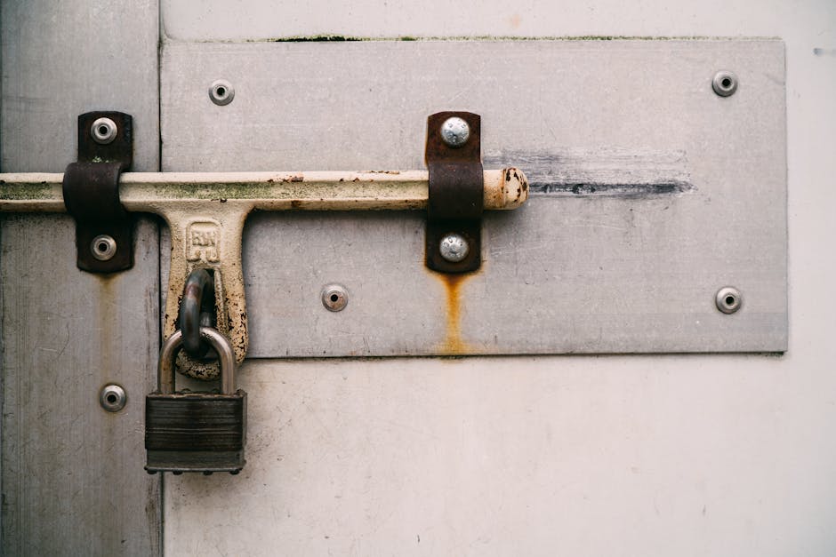Close-up image of a metal security latch and padlock affixed to a grey metal door, which appears weathered and slightly rusted, with visible bolt fastenings and rust stains underneath the latch. The l