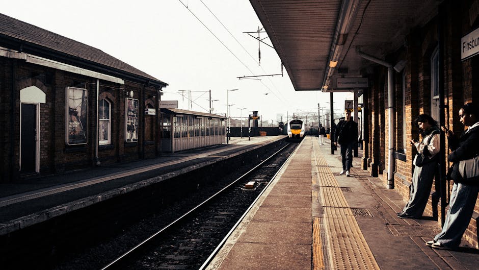 At Wanstead Park Station, a platform with a brick building on the right side features a row of three people standing and waiting, some holding bags or mobile devices. The platform is equipped with yellow tactile paving for visually impaired passengers, and a bench is visible beneath a shelter extending from the building. A yellow and white train approaches in the distance along railway tracks that run parallel to the platform, with overhead electrical wires and poles spanning across the tracks. On the left side, there is a separate platform with a building featuring large windows and advertisements, along with another train waiting. The scene is captured in natural daylight with overcast skies, emphasizing the station’s typical setting for home relocation or moving services. Man and Van Aldersbrook’s removals operations often involve using such stations for efficient collection and delivery of household furniture and packed boxes during packing and moving processes, demonstrating typical transportation and loading procedures in a railway environment.