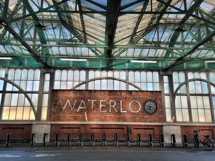 A street view outside Wanstead Park Station showing a large, historic brick building with arched windows and a sloped metal roof, situated above an underground station entrance. The foreground features a pavement area with a metal railing, a bicycle rack filled with bikes, and a covered bike shelter. Several people are standing or walking along the pavement, some carrying bags or engaged in conversation. Vehicles are parked along the street, and streetlights and trees with green foliage are visible on the right side of the image. The scene, taken during daylight hours with partly cloudy skies, captures a typical urban setting related to house removals and moving logistics, illustrating the environment where a professional removals company like Man and Van Aldersbrook might operate during a home relocation process.