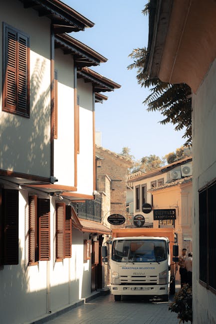 A narrow alleyway between two multi-storey buildings with white walls and brown wooden shutters on the windows. The alley is paved and a white van is parked at the end, with its front facing the camera. A person wearing casual clothing is standing near the van, possibly preparing for a home relocation or furniture transport. The scene is illuminated by natural daylight, with tree branches and foliage visible overhanging the building on the right. This setting illustrates a typical tight access situation for house removals, requiring careful planning and equipment, as managed by Man and Van Aldersbrook during packing and moving operations to ensure efficient loading and unloading within confined spaces.