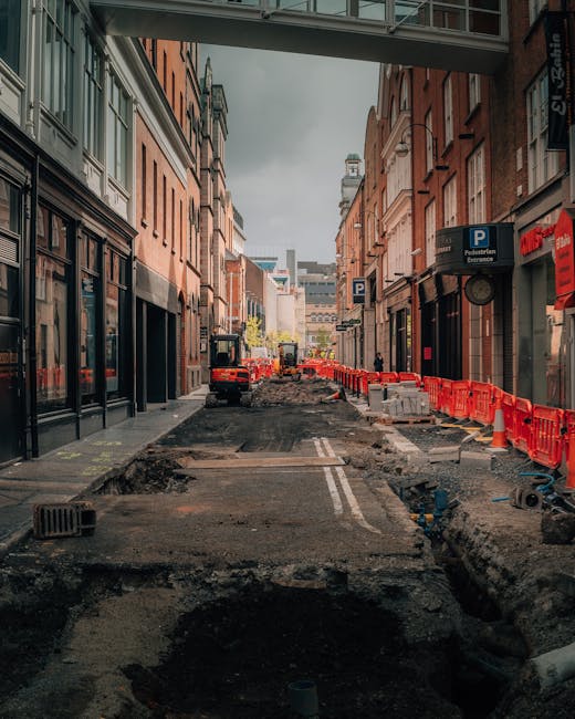 A city street undergoing road resurfacing with uneven, excavated asphalt surface, surrounded by multi-storey brick buildings on both sides. Roadwork barriers and construction signs line the area, with construction vehicles and machinery visible further down the street. The scene is captured from beneath a pedestrian bridge, with a partly cloudy sky overhead. This environment reflects the logistical challenges of house removals and furniture transport in areas with narrow or obstructed access, illustrating the types of logistical considerations that Man and Van Aldersbrook manages during home relocation projects involving disrupted roadways and confined spaces.