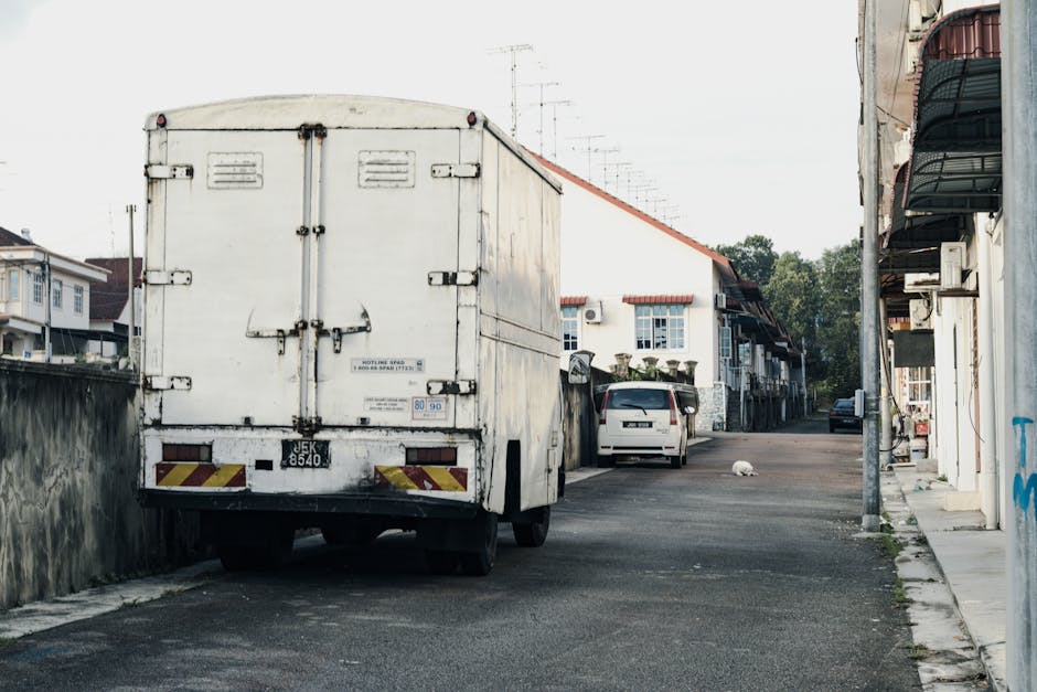 A narrow alleyway between two multi-storey buildings with white walls and brown wooden shutters on the windows. The alley is paved and a white van is parked at the end, with its front facing the camera. A person wearing casual clothing is standing near the van, possibly preparing for a home relocation or furniture transport. The scene is illuminated by natural daylight, with tree branches and foliage visible overhanging the building on the right. This setting illustrates a typical tight access situation for house removals, requiring careful planning and equipment, as managed by Man and Van Aldersbrook during packing and moving operations to ensure efficient loading and unloading within confined spaces.